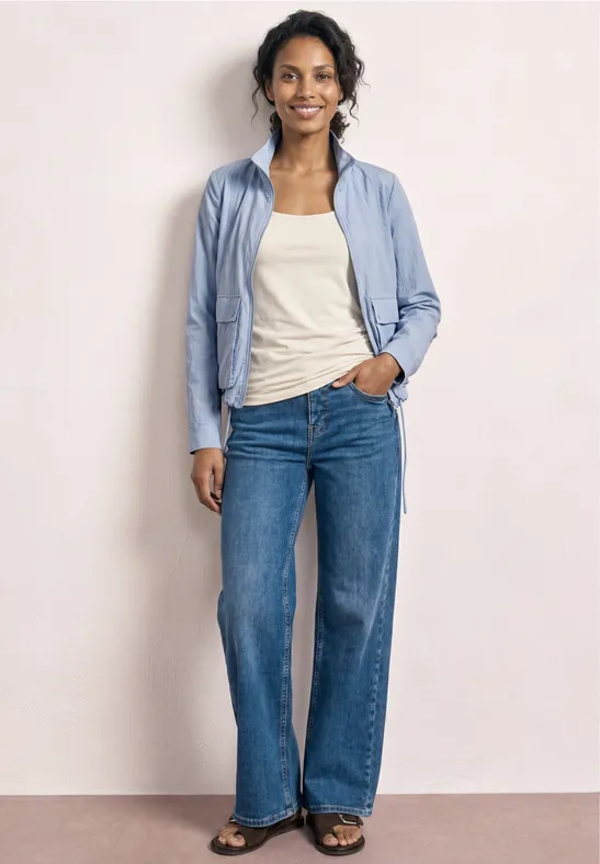 Femme devant un mur clair, chemise bleu clair, débardeur blanc et jeans larges bleu.