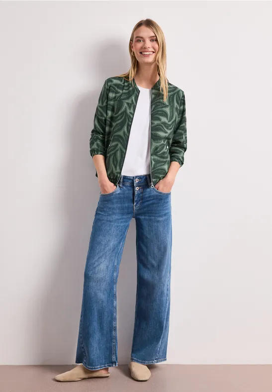 Frau in weißem T-Shirt, grüner Bomberjacke, weiten Jeans und beigen Schuhen, Hände in den Taschen.