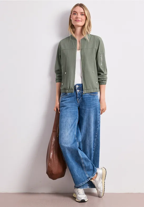 Person in grüner Jacke, weißem Top und weiten Jeans, mit brauner Tasche und Sneakern vor weißer Wand.