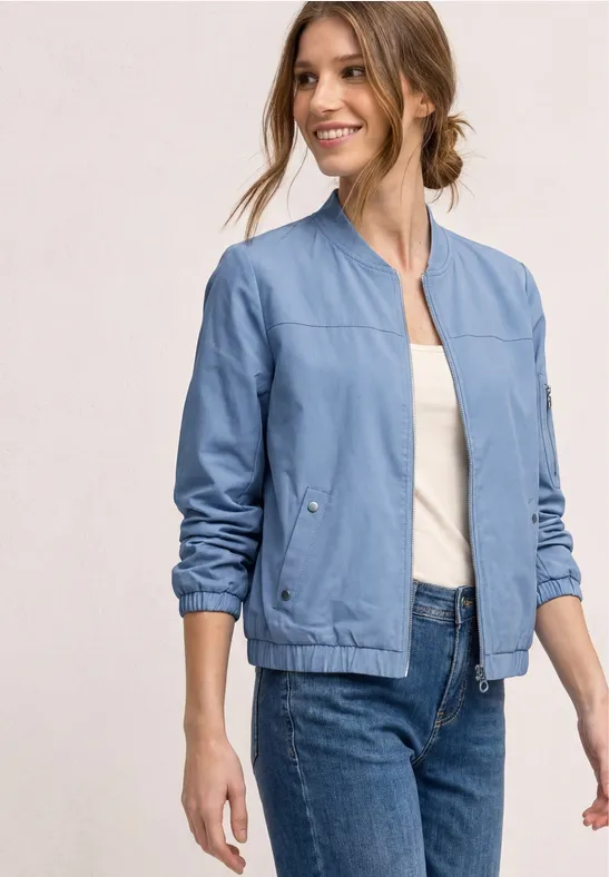 Een vrouw draagt een lichtblauw spijkerjack, wit hemd en blauwe jeans, gezicht vervaagd tegen lichte achtergrond.