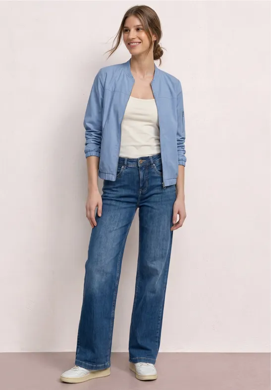 Een vrouw draagt een lichtblauw bomberjack, een wit topje, wijde high-waist jeans en witte sneakers.