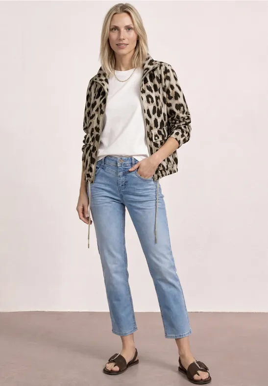Frau in Leopardenjacke, weißem T-Shirt, blauen Jeans und schwarzen Sandalen vor neutralem Hintergrund