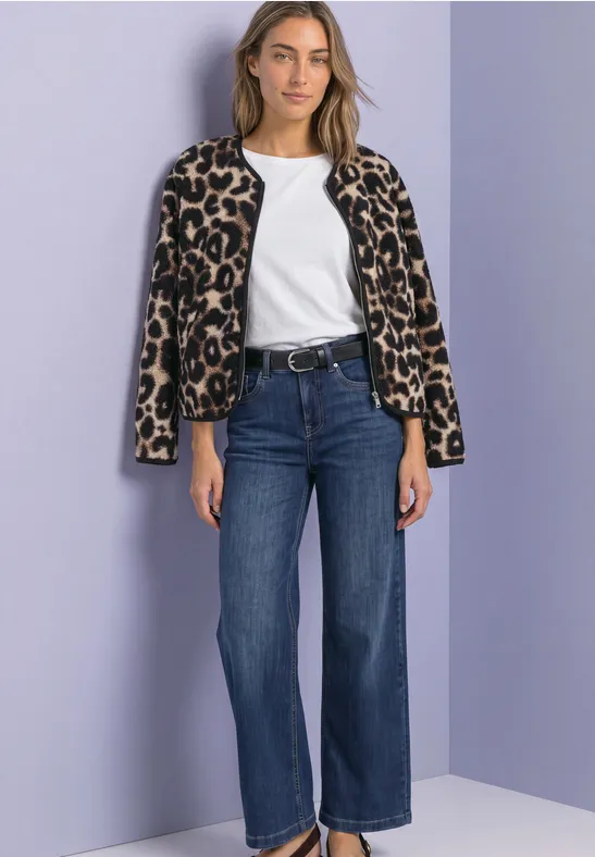 Eine Frau mit weißem Shirt, Leopardenjäckchen über den Schultern, blauen Jeans und Gürtel vor lila Wand.