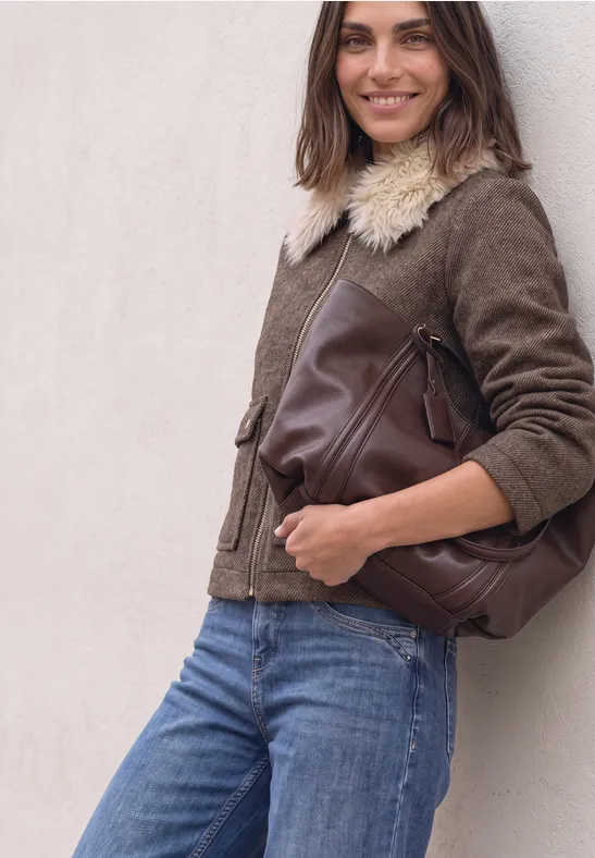 Frau lehnt lässig an Wand, trägt braune Jacke mit Fellkragen, große braune Ledertasche und blaue Jeans.