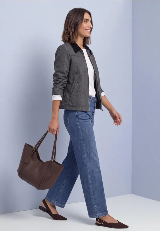 Frau in Jeans, weißem Shirt und grauer Jacke trägt eine braune Tasche und geht seitlich.