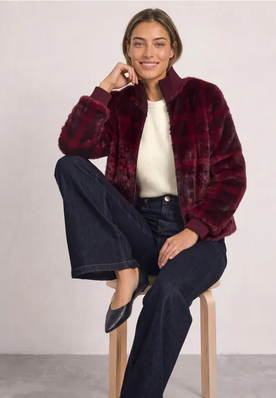 Frau in roter Plüschjacke, weißem Pullover und dunklen Jeans sitzt lässig auf einem Hocker vor grauem Hintergrund.