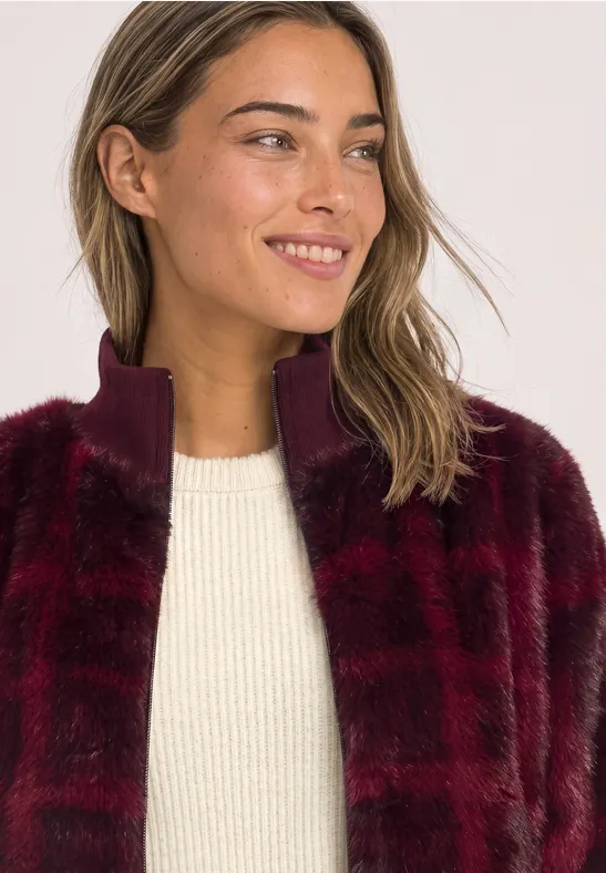 Eine lächelnde Frau mit weinroter Kunstfelljacke und weißem Pullover, Blick zur Seite.