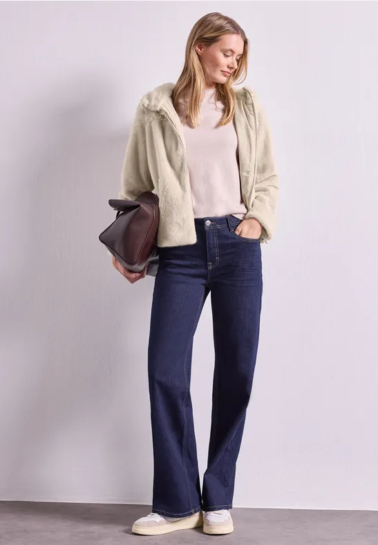 Frau in cremefarbener Fleecejacke, hellem Top, blauen Schlagjeans und weißen Sneakern, sie hält eine Tasche.