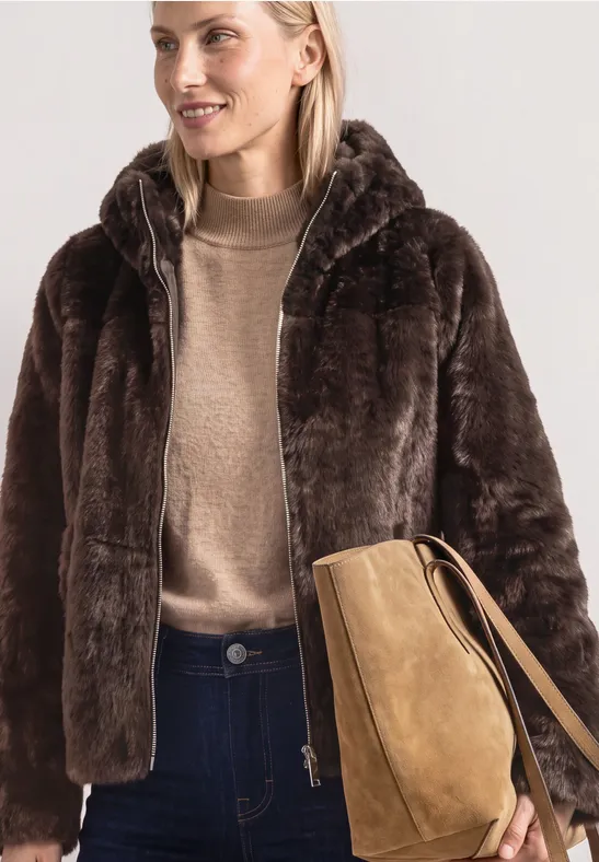 Frau in brauner Kunstfelljacke mit Kapuze, beigem Pulli, dunklen Jeans und camelfarbener Umhängetasche.