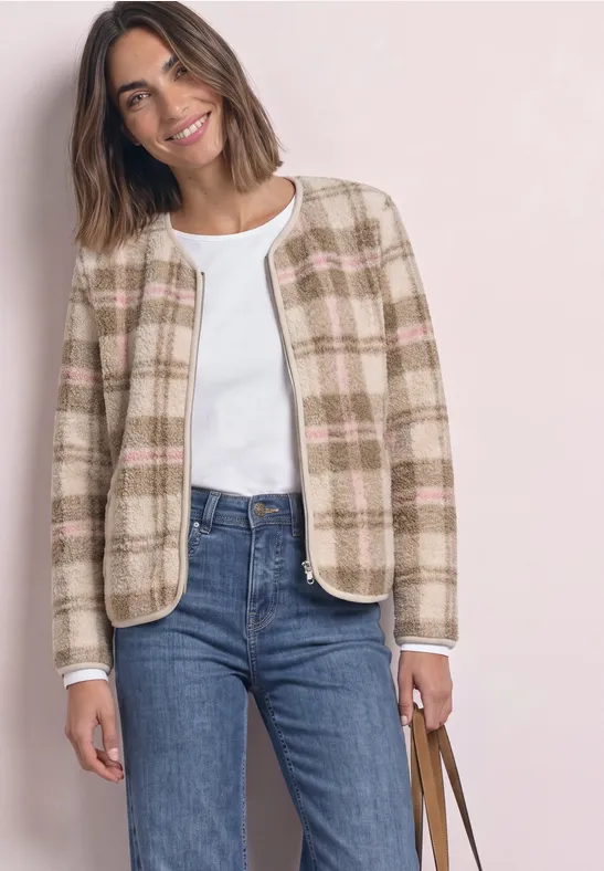 Frau in kariertem Kurzblazer, weißem Top, blauen Jeans und Tasche, lächelt.