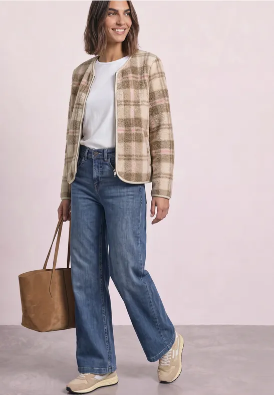 Frau in karierter beige Jacke, weißem T-Shirt, weiten Jeans, beigen Sneakern und brauner Tasche vor hellem Hintergrund.