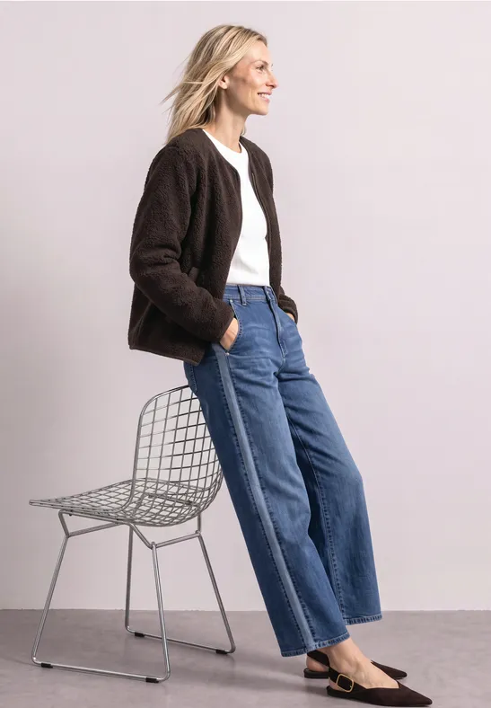 Frau in brauner Jacke, weißem Shirt, weiten Jeans und schwarzen Schuhen lehnt entspannt an einem Drahtstuhl.