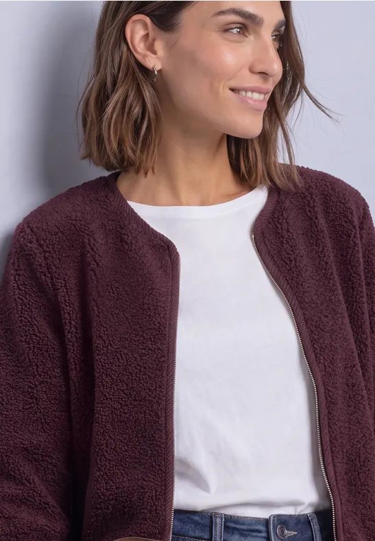 Frau in weißem T-Shirt und bordeauxfarbenem Teddy-Fleece-Jacke, lächelnd seitlich schauend