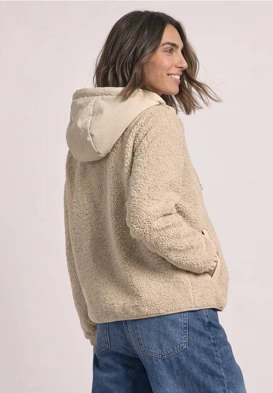 Frau in beiger Teddy-Fleece-Jacke mit Kapuze und blauer Jeans, seitlich lächelnd.