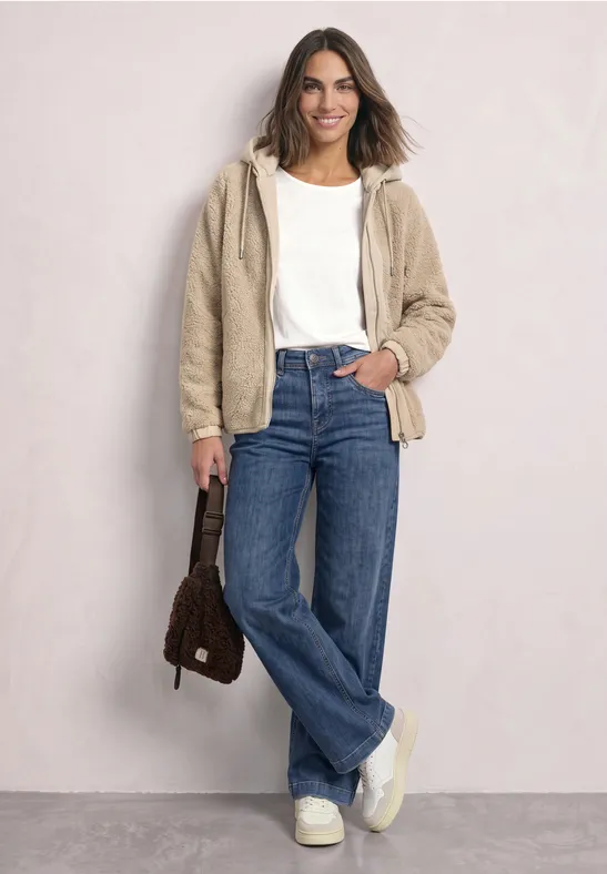 Frau in weißem Top, beiger Teddyjacke, Jeans und klobigen Sneakern, lächelnd, mit brauner Tasche.