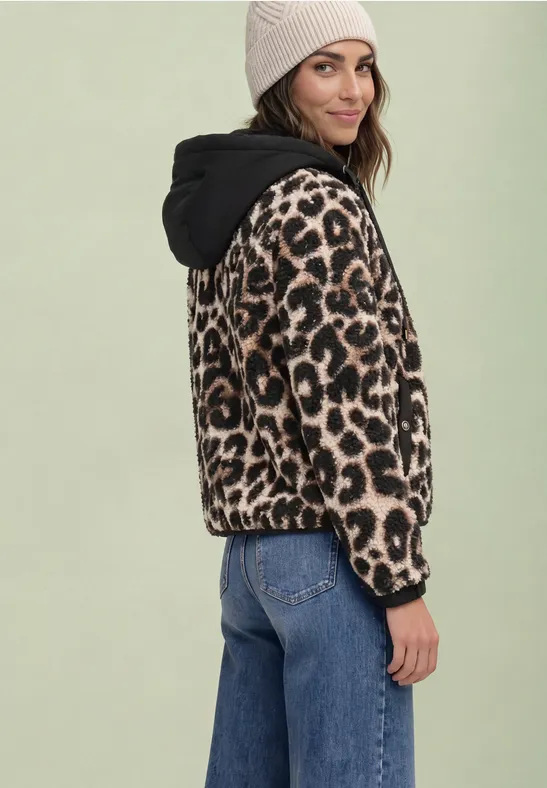 Frau in Leopardenjacke mit Kapuze, Beanie und Jeans, steht seitlich und lächelt.