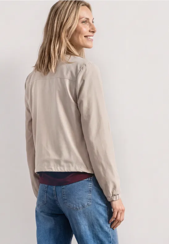 Eine Frau von hinten, sie trägt eine hellbeige Jacke und blaue Jeans vor hellem Hintergrund.
