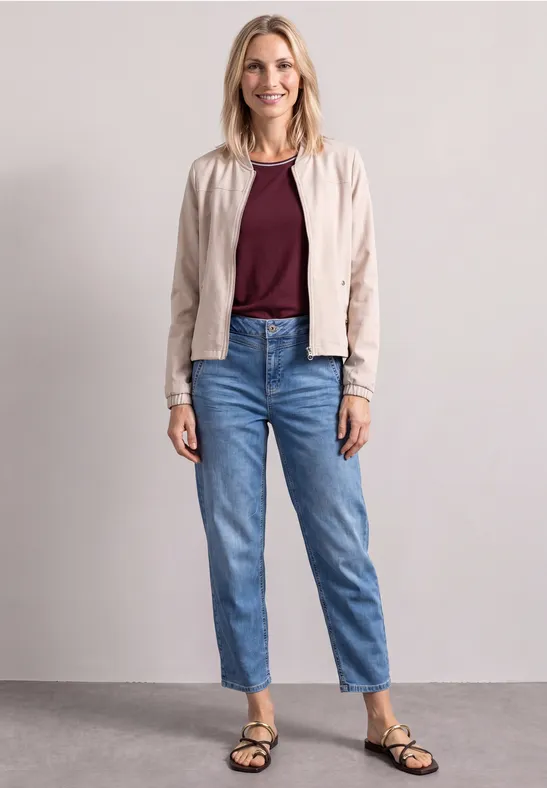 Frau in Jeans, bordeauxrotem Top, heller Jacke und Sandalen vor neutralem Hintergrund.