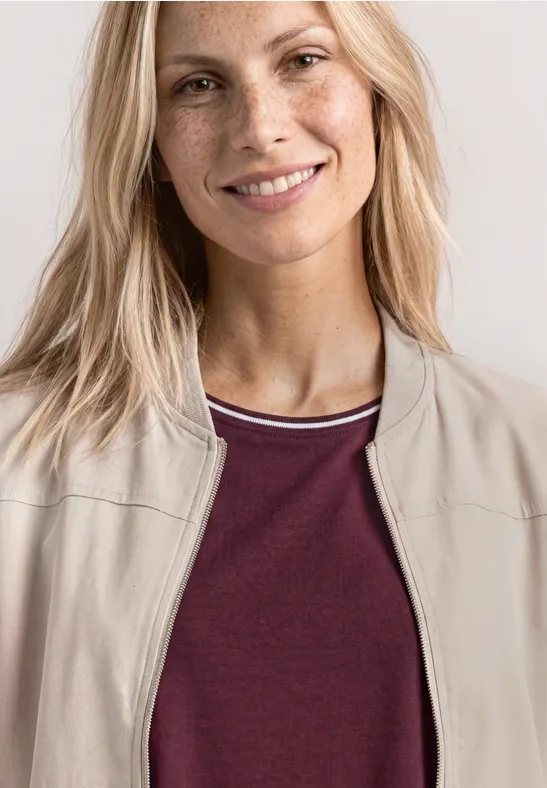 Blonde Person von den Schultern aufwärts, trägt eine beige Jacke und ein bordeauxrotes Shirt.