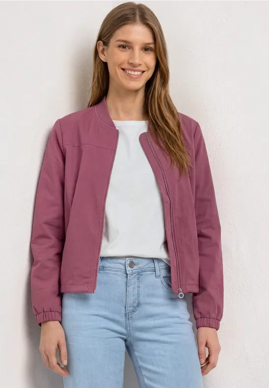 Een vrouw draagt een paarse korte jas over een wit T-shirt en spijkerbroek, tegen een lichte achtergrond.