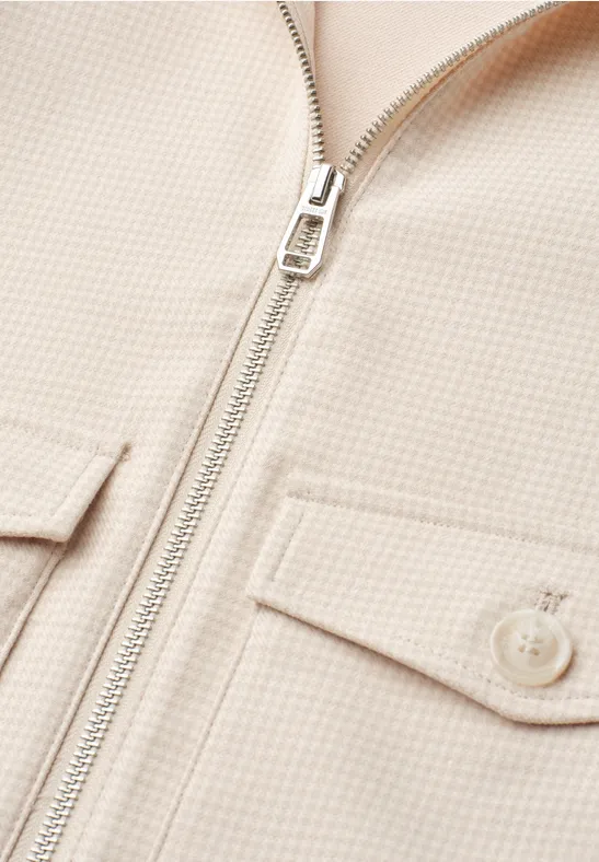 Vue rapprochée d'une veste beige avec fermeture éclair métallique et deux poches poitrine à rabat.