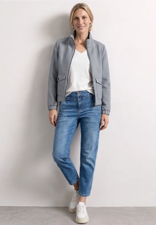 Femme en t-shirt blanc, blazer gris, jean bleu et baskets blanches, posant devant un mur clair.