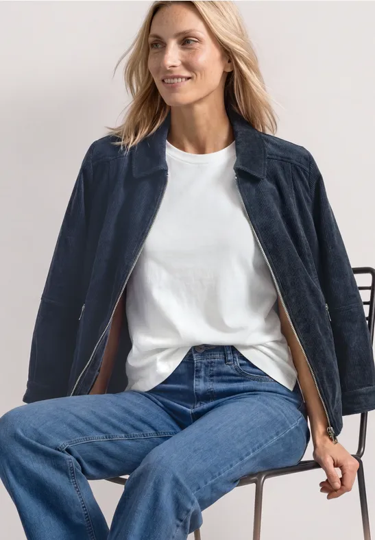 Frau in weißem T-Shirt und Jeans sitzt auf Stuhl, trägt dunkelblaue Jacke.