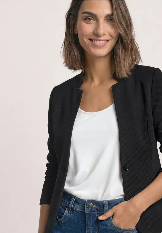Eine lächelnde Frau in schwarzem Blazer und weißem Shirt, Hand in der Hosentasche.