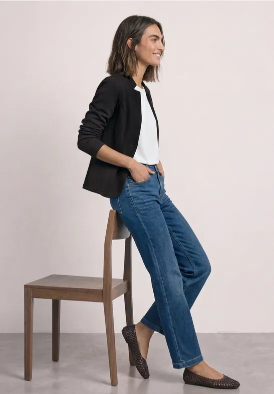 Frau in legerem Outfit: schwarzer Blazer, weißes Shirt, blaue Jeans und flache Schuhe lehnt an einem Stuhl.