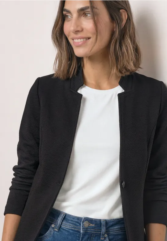 Frau in schwarzer Jacke und weißem Shirt, lächelnd vor hellem Hintergrund.