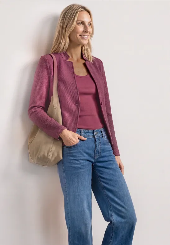 Vrouw draagt frambozenkleurig jasje, bijpassende top, blauwe jeans en beige stoffen tas voor witte muur.