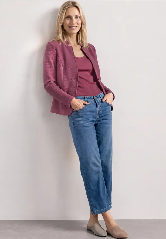 Vrouw leunt tegen witte muur, draagt paarse blazer en top, blauwe jeans en beige ballerina's.