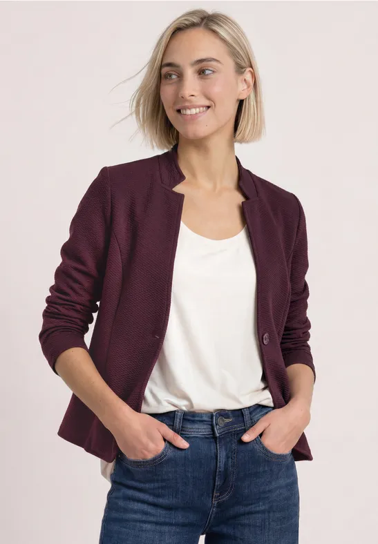 Eine lächelnde Frau im weißen Top, burgunderrotem Blazer und blauen Jeans posiert mit Händen in den Hosentaschen.