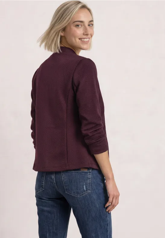 Frau mit blondem Haar in burgunderrotem Blazer und Jeans schaut über ihre Schulter.