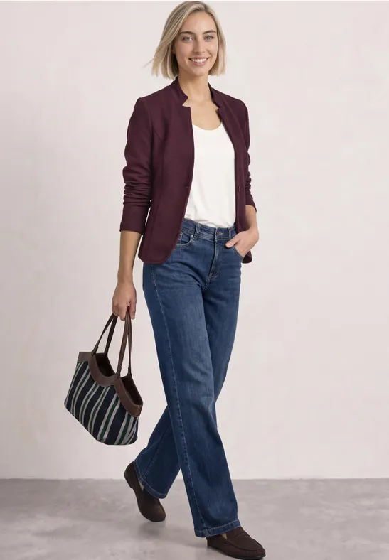 Eine Frau in weißem Top, bordeauxrotem Blazer und blauen Jeans hält eine gestreifte Tasche und lächelt.