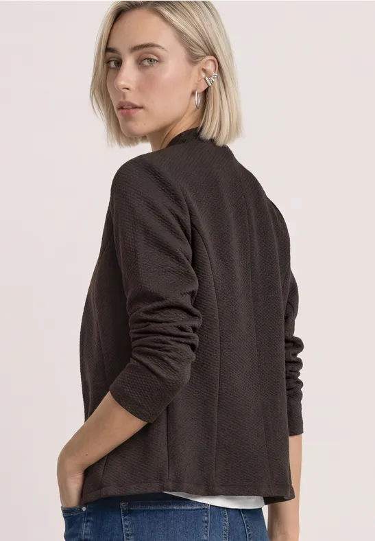 Frau mit kurzem blondem Haar trägt einen schwarzen Blazer, steht seitlich und blickt über die Schulter.