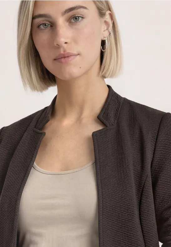 Frau mit kurzem blondem Haar trägt eine braune Jacke und ein beiges Top, Blick ruhig und direkt.
