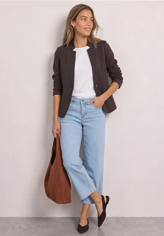 Frau in braunem Blazer, weißem Shirt und hellblauen Jeans, trägt große braune Tasche und schwarze Ballerinas.