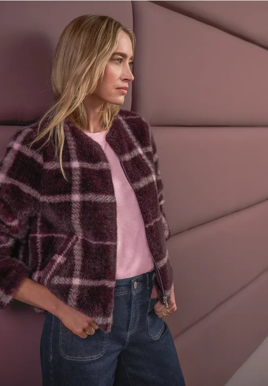 Dame mit kariertem Mantel lehnt an gepolsterter Wand, trägt rosa Shirt und Jeans.
