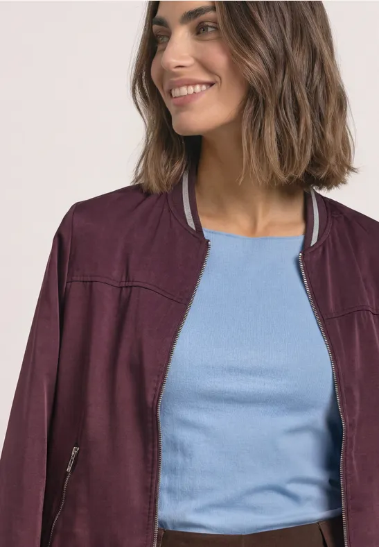 Eine Frau trägt eine violette Bomberjacke und ein hellblaues Shirt, leichtes Lächeln.