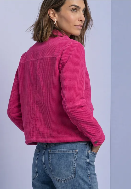 Femme en veste rose et jean bleu regarde par-dessus son épaule sur fond clair.