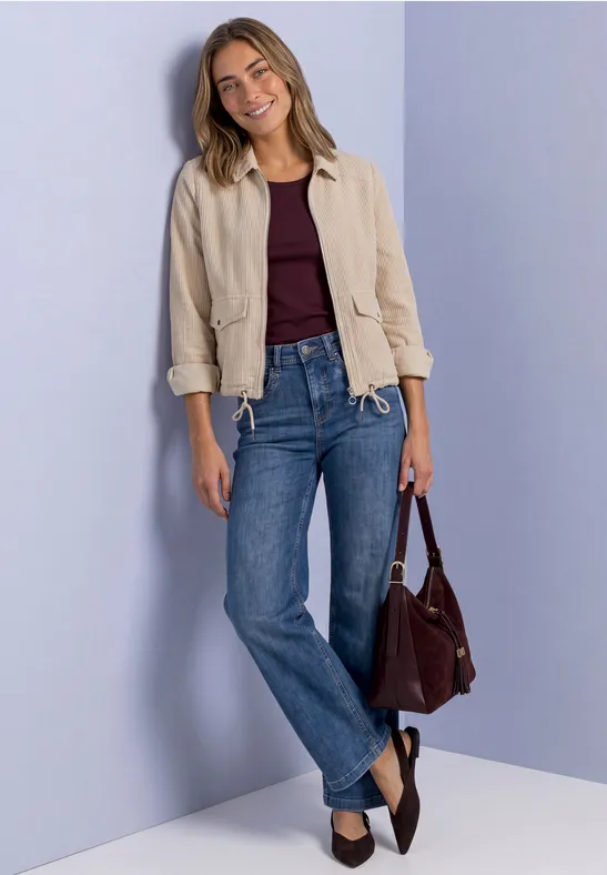 Junge Frau in Jeans, bordeauxrotem Top und heller Jacke trägt eine braune Handtasche vor helllila Hintergrund.