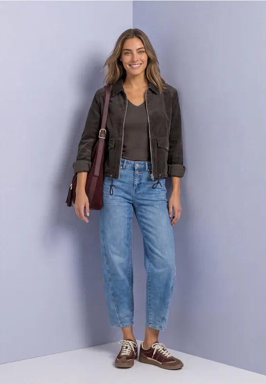Frau in Lederjacke, schwarzem Top, Jeans und Bootsschuhen lächelt und trägt eine Umhängetasche vor fliederfarbener Wand.