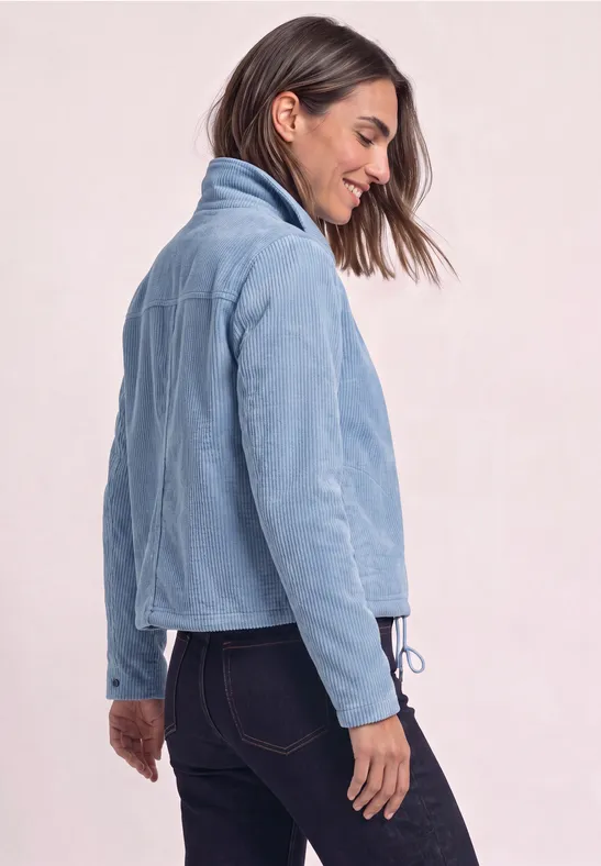 Femme de profil portant une veste en velours côtelé bleu clair et un jean foncé sur fond neutre.