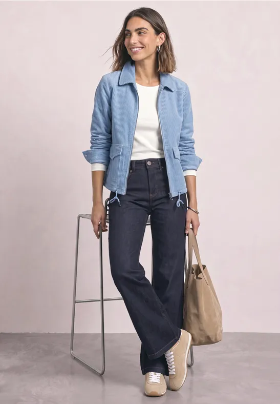 Femme en veste bleue, haut blanc, jean évasé et chaussures claires, porte un sac beige sur fond neutre.