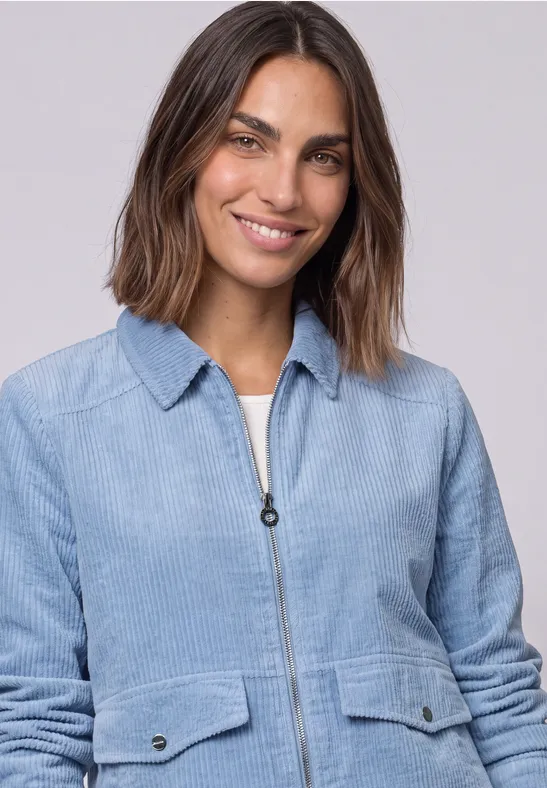 Femme aux cheveux bruns mi-longs porte une veste en velours côtelé bleu clair et sourit à la caméra.