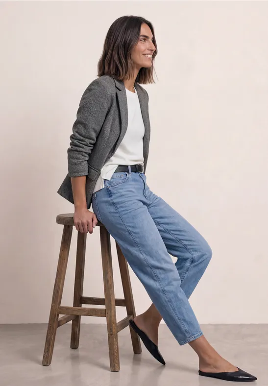 Frau sitzt entspannt auf einem Hocker, trägt grauen Blazer, weißes Top, blaue Jeans und flache schwarze Schuhe.
