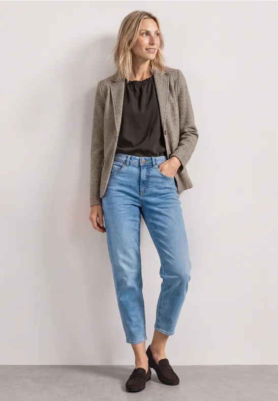 Kobieta z blond włosami ma na sobie szarą marynarkę, czarny t-shirt, niebieskie jeansy i czarne mokasyny.