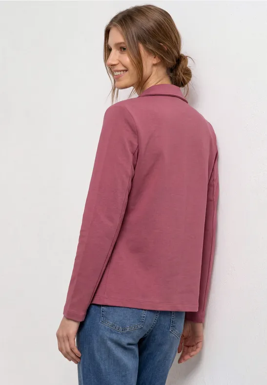 Eine Frau trägt eine rosafarbene langärmlige Jacke mit Reverskragen und Jeans, blickt über die Schulter.