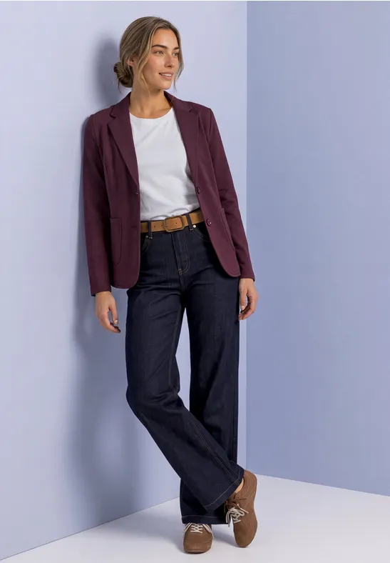 Vrouw leunt tegen een pastelmuur, gekleed in wit T-shirt, donkerblauwe jeans, bordeauxblazer en bruine schoenen.
